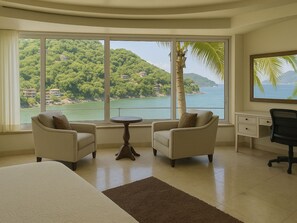 A hotel room with a large window offering a view of a lush green hillside and a body of water. There is a bed, two armchairs, a small round table, and a desk with a chair. - Ferienvilla "Villa Armonia" mit Infinity Pool by Interhome (Boca de Tomatlan)