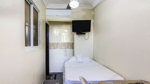 Una habitación de hotel estrecha con cama, televisor montado en la pared y ventilador de techo.