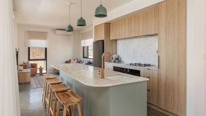 A modern kitchen with wooden cabinets, a central island with stools, pendant lighting, and a built-in dishwasher. - ESCAPE TO PICTURESQUE EAGLE BAY IN THIS BEAUTIFULLY APPOINTED 2 BED STUDIO (Eagle Bay)