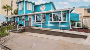 Une maison de plage turquoise avec une enseigne indiquant « CaliMyths Beach Resort ». Elle possède une balustrade blanche, un escalier et offre une vue sur l’océan.