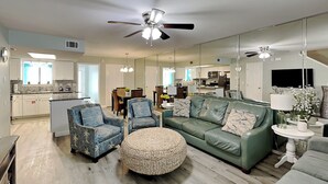 Un salon moderne avec un canapé vert, deux fauteuils, un pouf rond et un coin repas visible à travers un grand miroir.