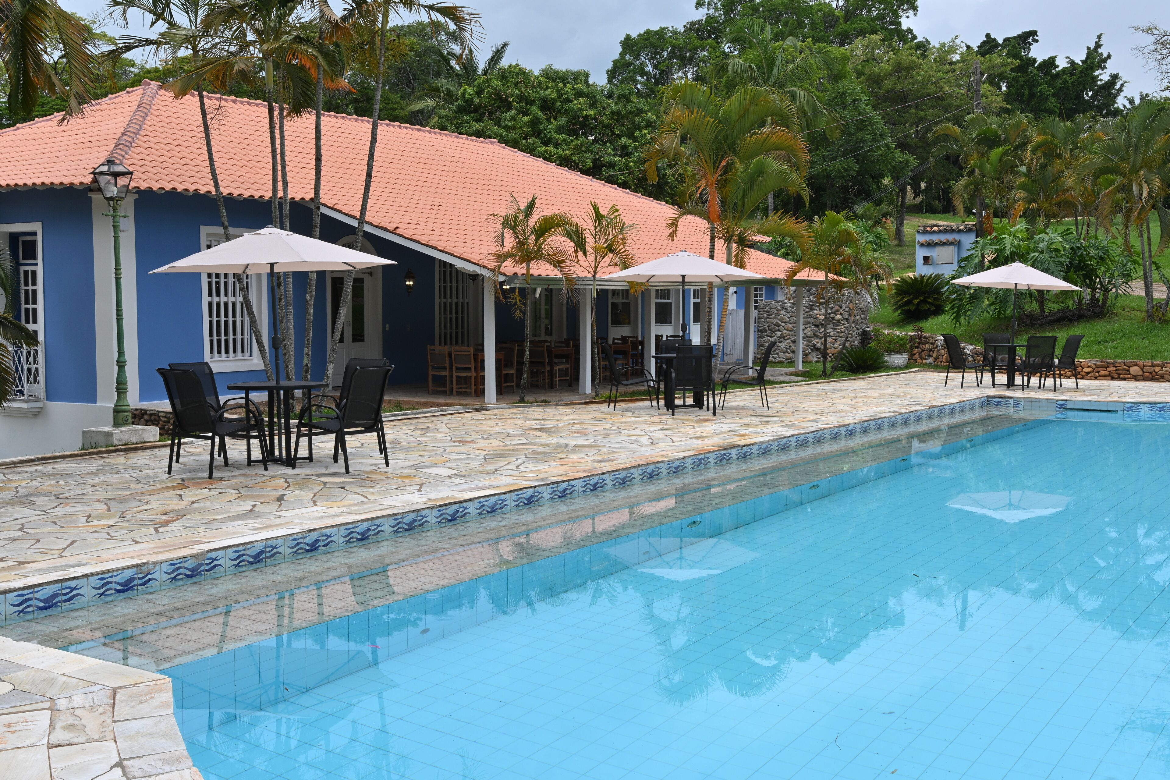 A pool area with a tiled edge, outdoor seating, and a building with a red-tiled roof.