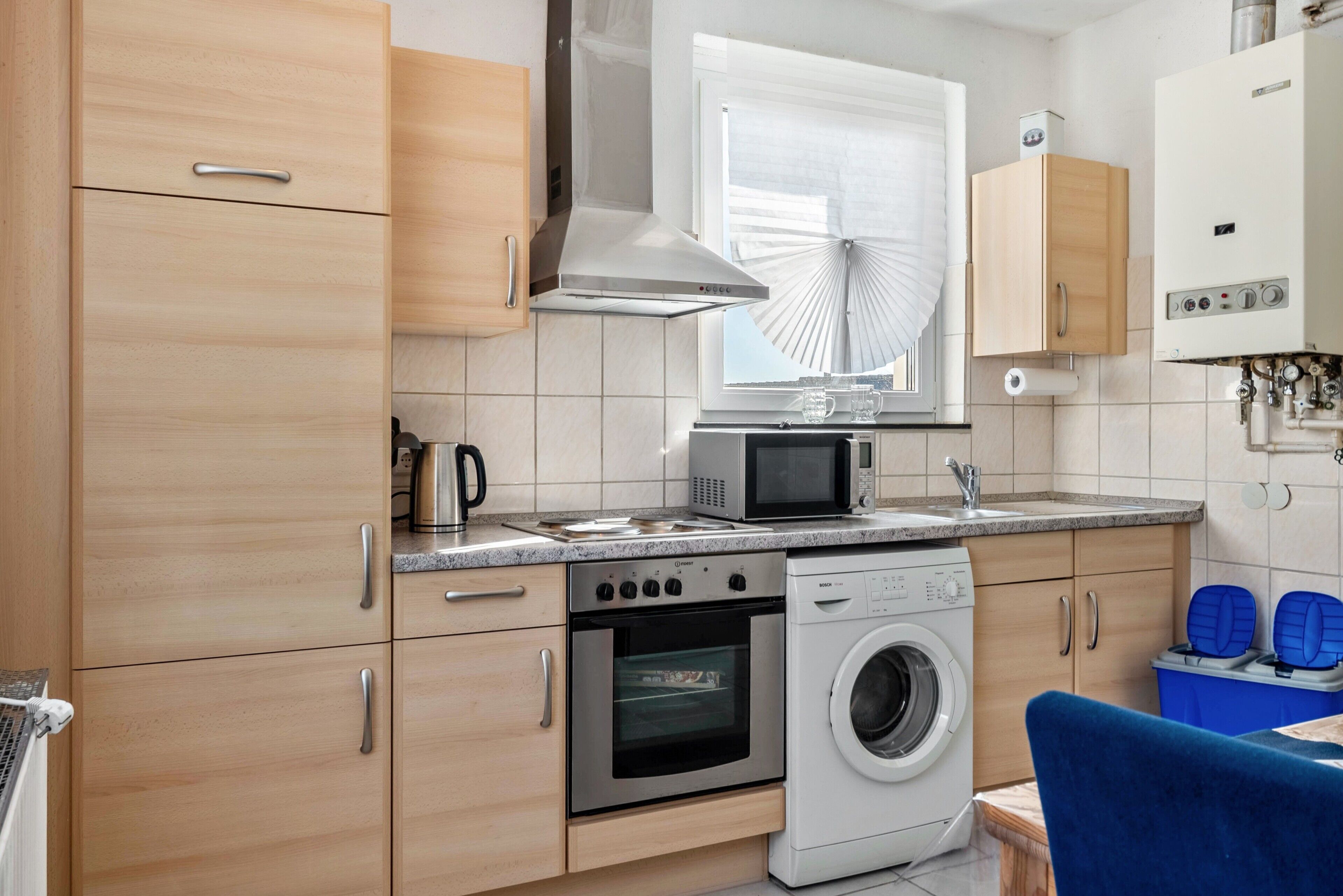 A compact kitchen with a microwave, oven, and washing machine, wooden cabinets, and a window with blinds.