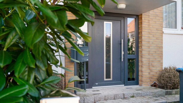 Una entrada moderna de edificio con puerta oscura, escalones y un pequeño jardín.