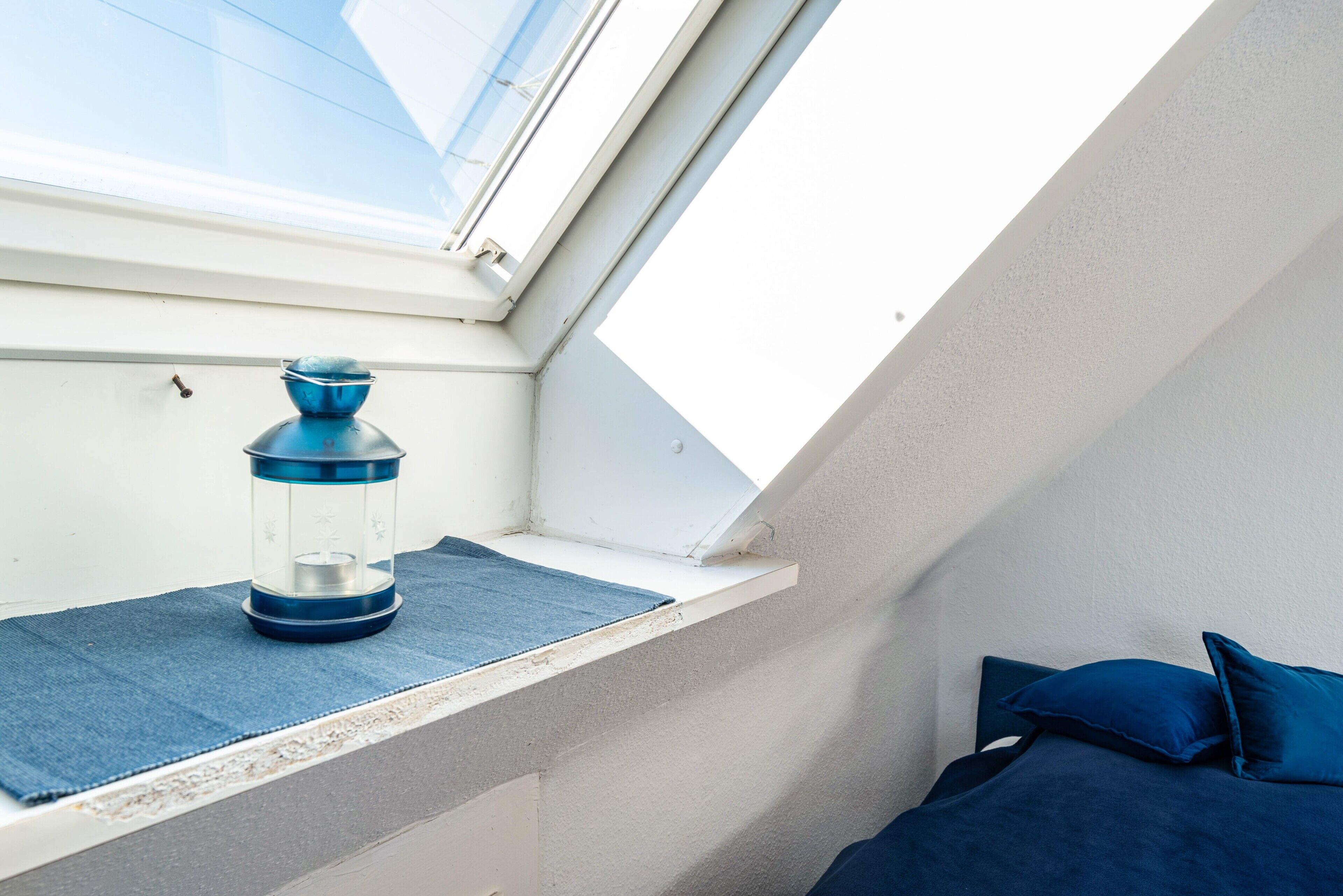 Een dakraam met een blauwe lantaarn op de vensterbank, een bed met blauw beddengoed en een witte muur.