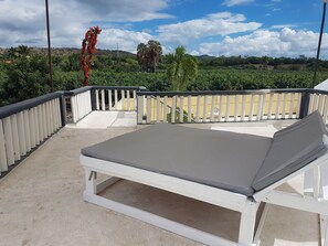 A sunbed on a balcony with a white railing, overlooking a landscaped garden with palm trees and a view of distant hills.