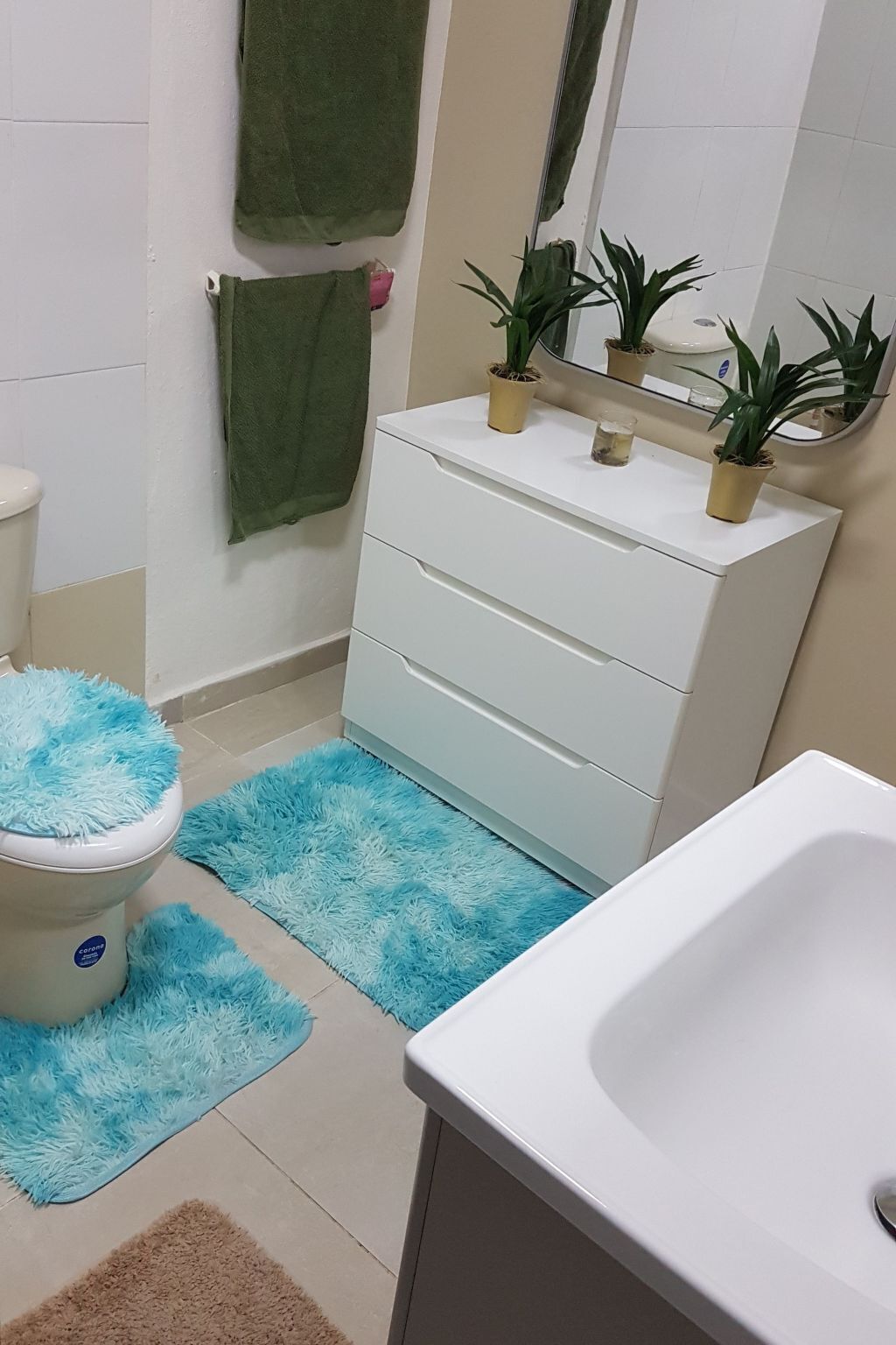 Une salle de bain avec un lavabo blanc, des toilettes dotées d’un abattant bleu et moelleux, un meuble-lavabo blanc avec trois tiroirs, et deux plantes en pot posées sur le meuble-lavabo.