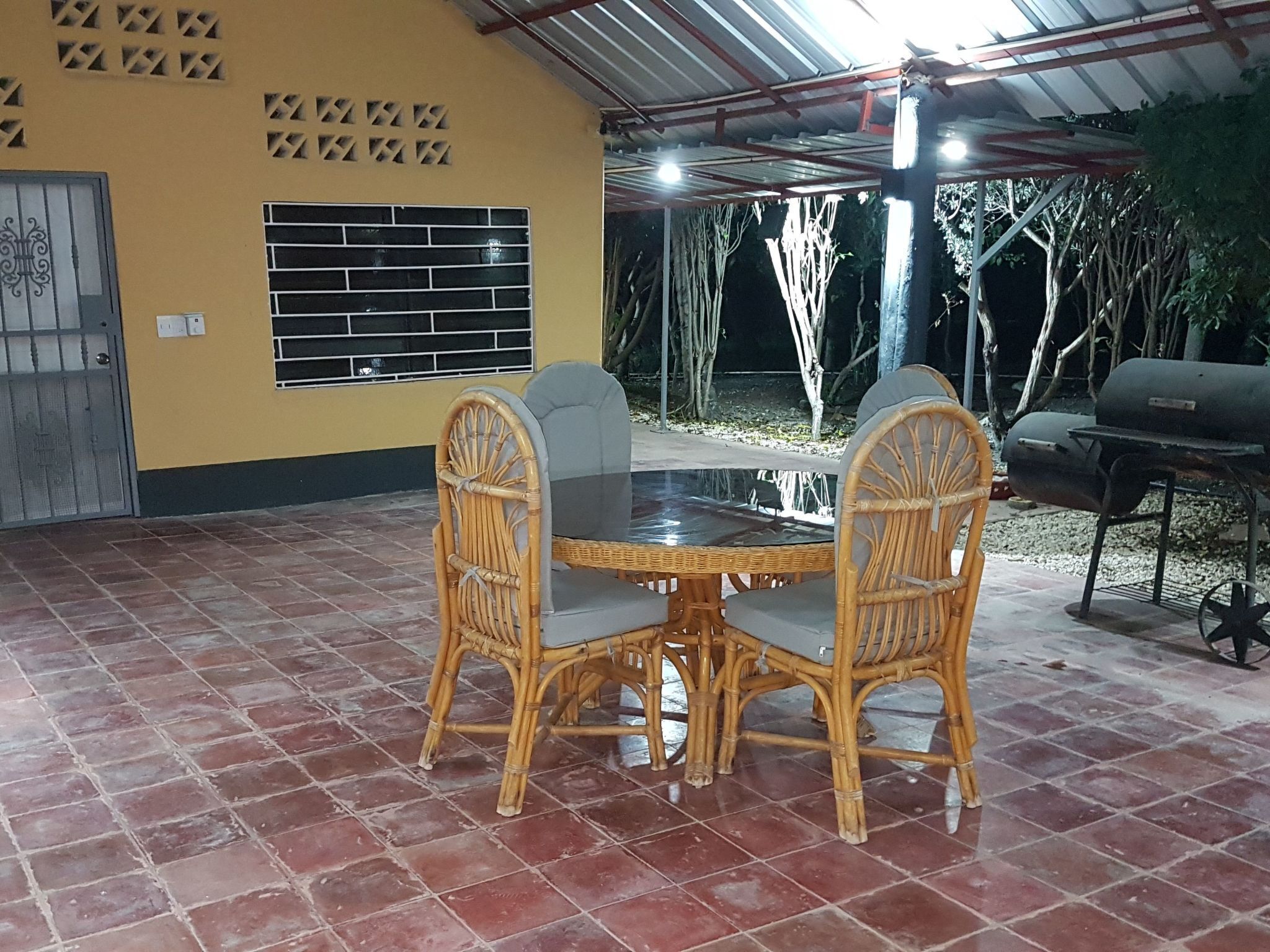 Un patio al aire libre con una mesa redonda y cuatro sillas de mimbre, una parrilla y un área techada con techo metálico.