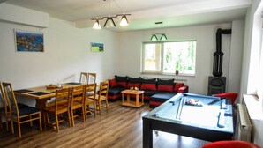 Una sala de estar con mesa de billar, una mesa de comedor de madera y sillas. Hay un sofá con cojines rojos, una ventana con vista a la vegetación y una lámpara de techo.