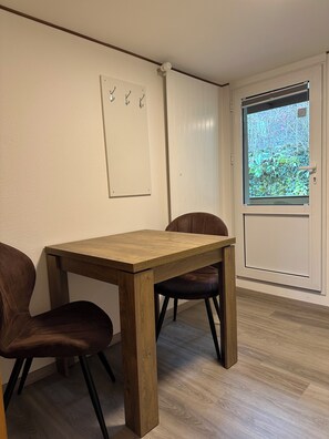 Un coin repas avec une table en bois et deux chaises, une porte blanche avec une fenêtre et un panneau mural avec des crochets.