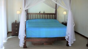 Ein Bett mit Baldachin, ausgestattet mit einer blauen Bettdecke und einem weißen Moschee-Vorhang, steht zwischen Nachttischen mit Lampen, in einem Raum mit schlichten Wänden und Fliesenboden.