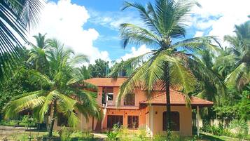 A two-story house with a red-tiled roof, surrounded by lush greenery and palm trees.