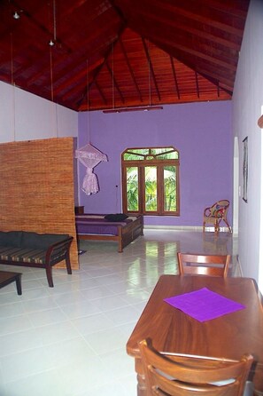 A spacious room with a high ceiling, a wooden dining table with chairs, a bed with a purple bedspread, a hanging chair, and a window with a view of greenery. - Peacock by Interhome (Habaraduwa /Galle)