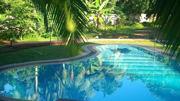 A swimming pool surrounded by lush greenery and palm trees, with a clear blue water surface reflecting the surrounding vegetation.