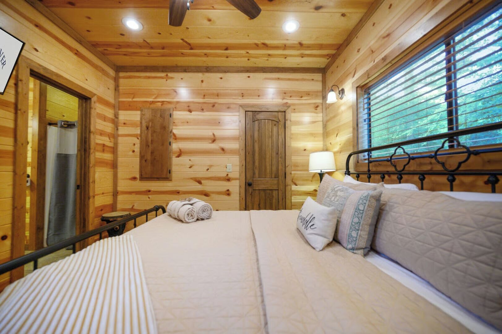 A wooden cabin bedroom with a bed, pillows, a nightstand, a window with blinds, and a door.