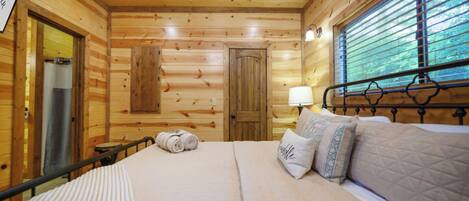 A wooden cabin bedroom with a bed, pillows, a nightstand, a window with blinds, and a door.