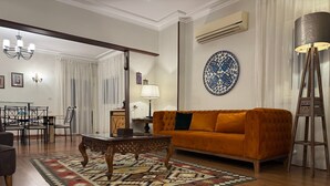 Ein Wohnzimmer mit Sofa, Couchtisch und einem gemusterten Teppich. Im Hintergrund befindet sich ein Essbereich mit Stühlen und Tisch.