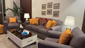Ein Wohnzimmer mit einem grauen Sofa, zwei Sesseln, einem Couchtisch und einer Lampe. An den Wänden hängen Bilder in Rahmen und es steht ein Blumentopf dort.