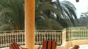 A covered outdoor seating area with wicker chairs and a table, surrounded by a white balustrade and palm trees in the background.