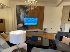 A modern living room with a flat-screen TV mounted on a marble wall, a chandelier, and a sofa set. - "Classic Style apart". by Interhome (Abu Al Feda)