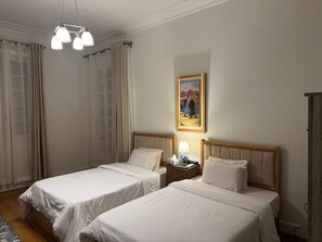 A hotel room with two single beds, a nightstand with a lamp, a painting on the wall, and a wooden wardrobe. - "Classic Style apart". by Interhome (Abu Al Feda)