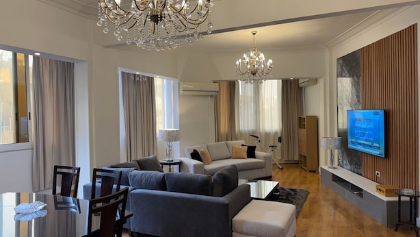 Ein modernes Wohnzimmer mit einer großen Kronleuchter, einem grauen Sofa, einem Couchtisch und einem an der Wand montierten Fernseher.