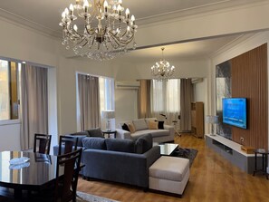 Ein modernes Wohnzimmer mit einer großen Kronleuchter, einem grauen Sofa, einem Couchtisch und einem an der Wand montierten Fernseher.
