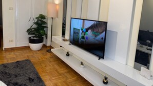 A modern living room with a flat-screen TV mounted on a white console, a potted plant, and a floor lamp. - "Gemütliche 1-Zimmer-Wohnung". by Interhome (Abu Al Feda)