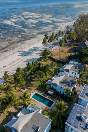 Vista aérea de un complejo turístico frente al mar, con varias edificaciones, una piscina y palmeras.