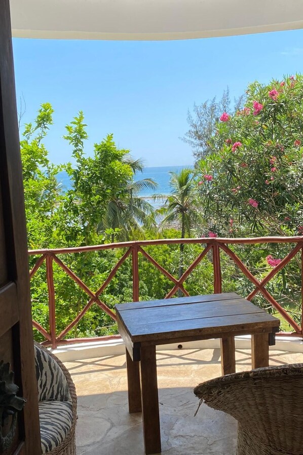 A balcony with a table and wicker furniture, overlooking a tropical landscape with trees and a view of the sea.