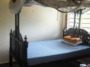 Un lit simple avec un matelas bleu et une tête de lit en bois, une petite table avec un coussin et une fenêtre avec des rideaux.