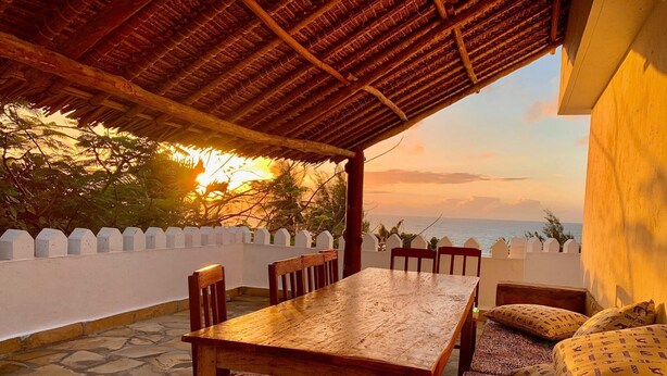 Eine Terrasses mit Holz-Tisch und -Stühlen, einem Reetdach und Blick auf das Meer bei Sonnenuntergang.