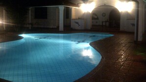 Una piscina con bordo piastrellato, un'area relax coperta e un edificio con finestre ad arco sullo sfondo.