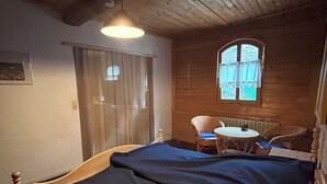 Una habitación acogedora con techo de madera, una cama con cubierta azul, una mesita redonda y un ventanal con cortinas.