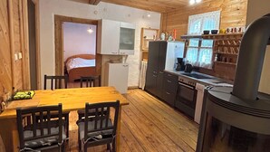 Interior acogedor de una cabaña con una mesa de madera, sillas, una zona de cocina con estufa y una cama al fondo.