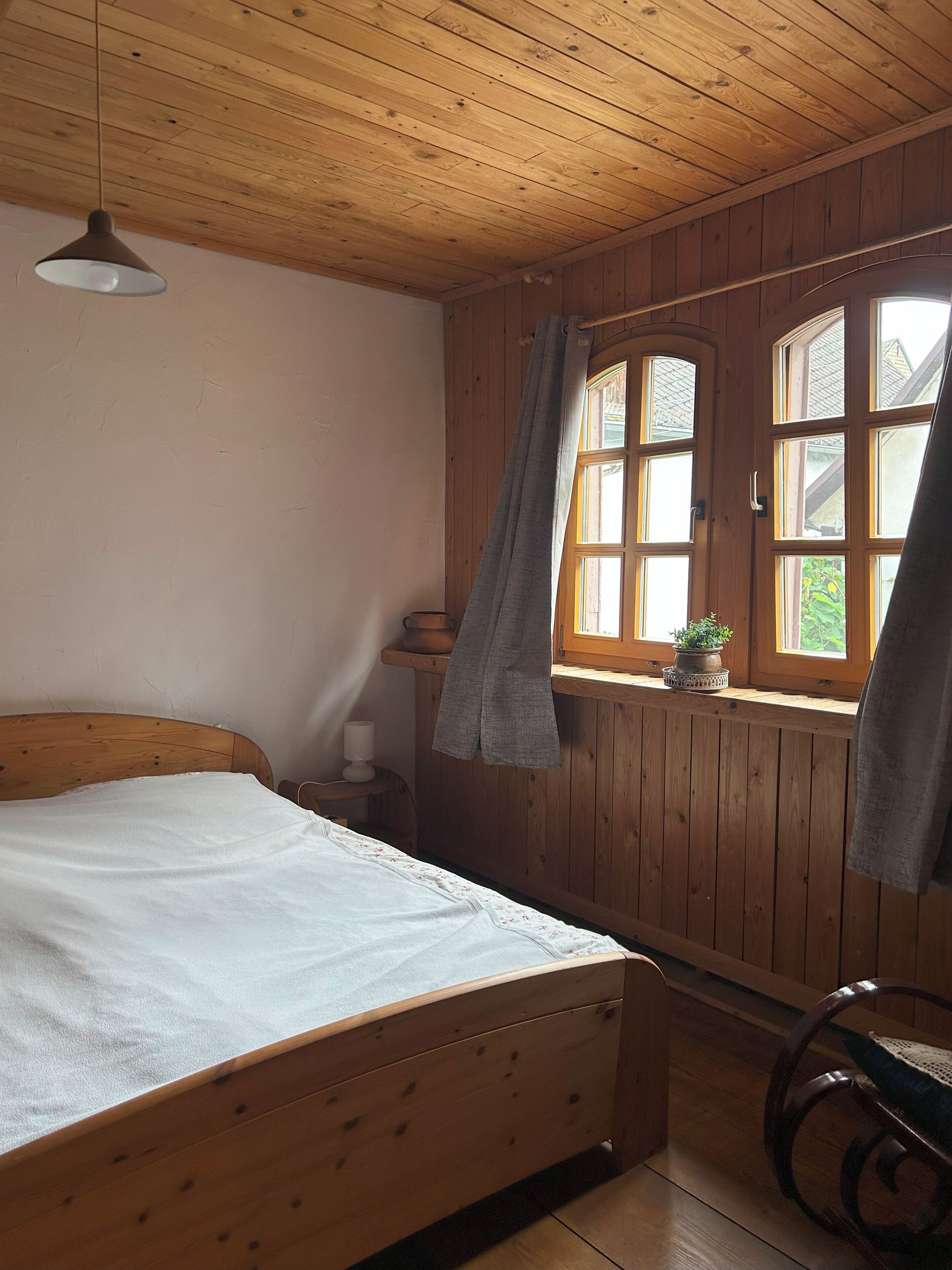Ein Zimmer mit Holzwänden, einem Bett, einem Fenster mit Vorhängen, einem kleinen Tisch und einem Stuhl.