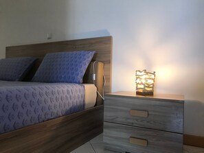 Una cama con colcha estampada y almohadas, una mesita de noche con lámpara y una cómoda de madera con cajones.