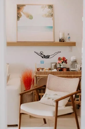 A room with a white sofa, a wicker chair, a wall-mounted shelf with decorative items, and a framed artwork depicting a swimmer. - Strandparadies Pur by Interhome (500-Cumbuco)