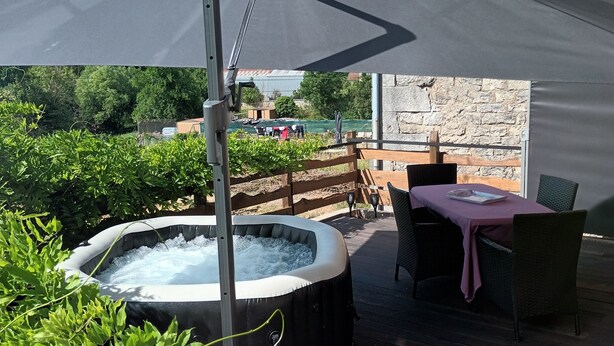 An outdoor hot tub area with a table and chairs under a large umbrella, surrounded by greenery and a stone wall. - Country house with jacuzzi and pets allowed (Saint-Joire)