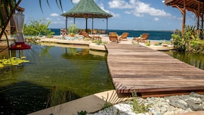 Una terraza de madera que conduce a un gazebo con techo verde, con vistas a una masa de agua tranquila y el océano.