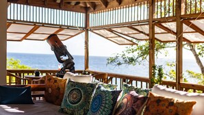 Una terraza de madera con una cama, almohadas estampadas, un telescopio y vista al océano.