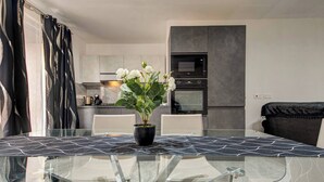 Une cuisine moderne dotée d’un four et d’un micro-ondes encastrés, un coin repas avec une table et des chaises en verre, et une plante en pot ornée de fleurs blanches.
