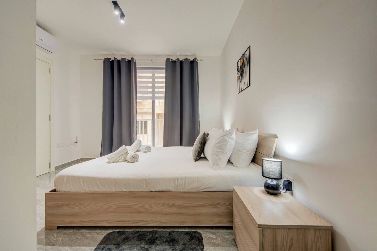 A modern bedroom with a wooden bed, grey curtains, a bedside lamp, and a view of the outside balcony.