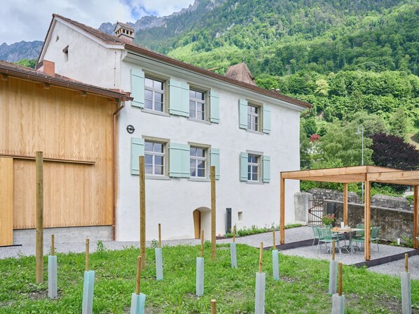 Une maison à deux étages aux murs de couleur claire et aux volets verts, agrémentée d’une pergola en bois et d’un jardin planté de jeunes arbres.