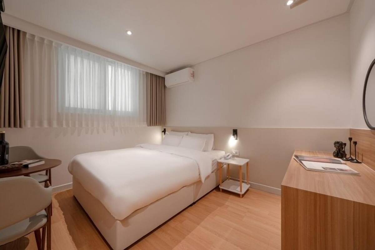 A hotel room with a bed, bedside table, desk, chair, and a window with curtains.