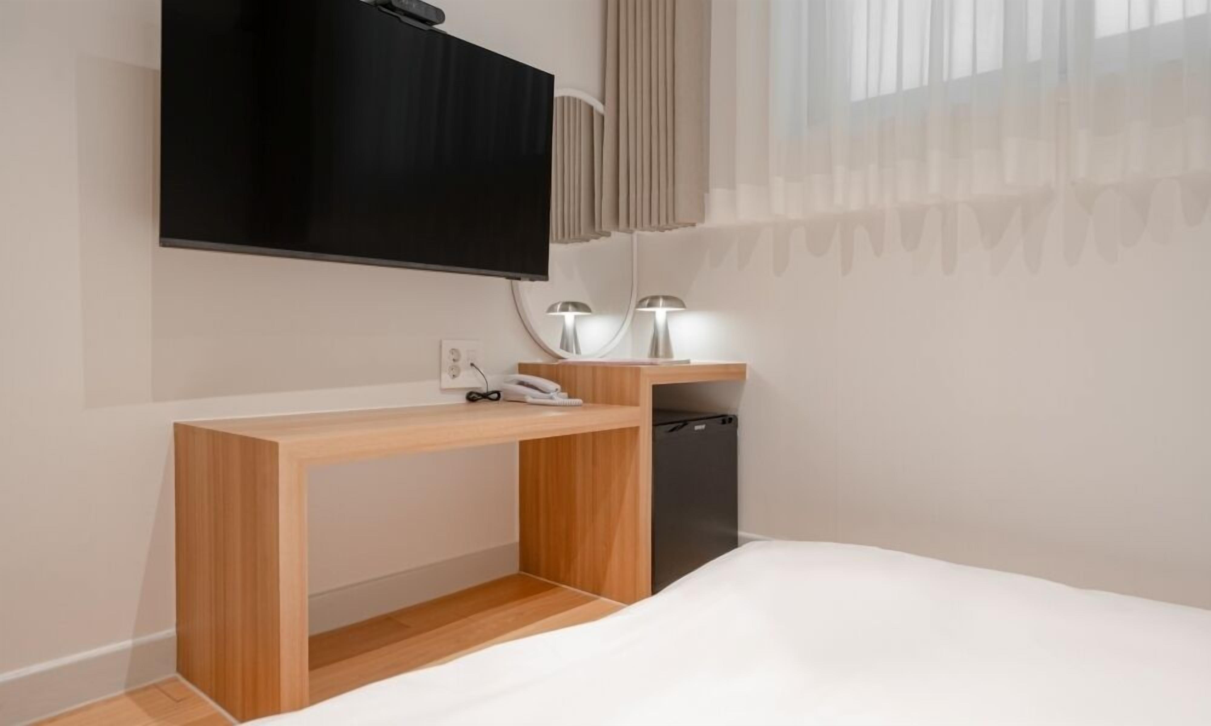 Habitació d'hotel moderna amb escriptori de fusta, televisió de pantalla plana penjada a la paret, taula de nit petita amb làmpades i llit amb roba de llit blanca.
