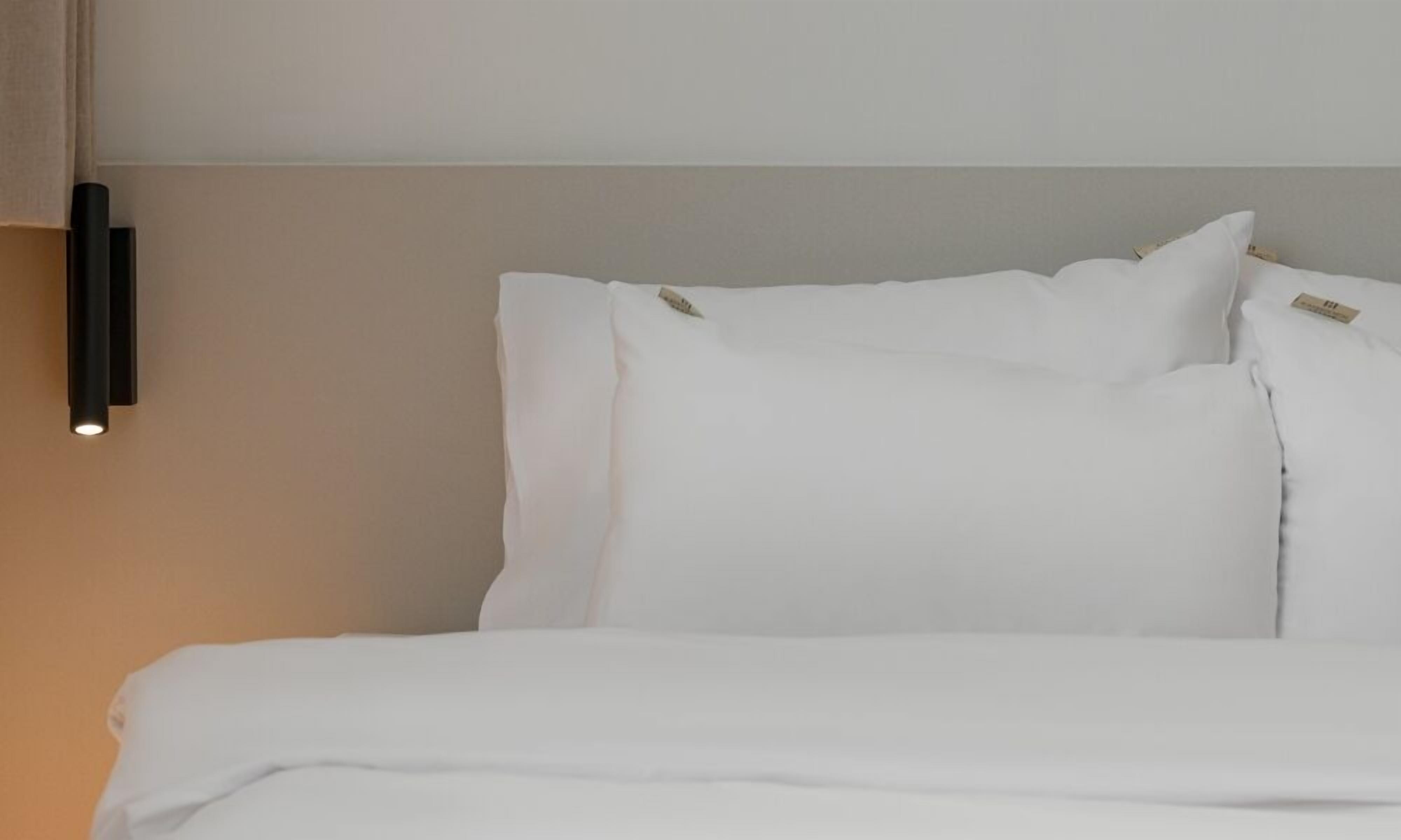 A neatly made bed with white bedding and pillows, a wall-mounted light fixture, and a plain wall.