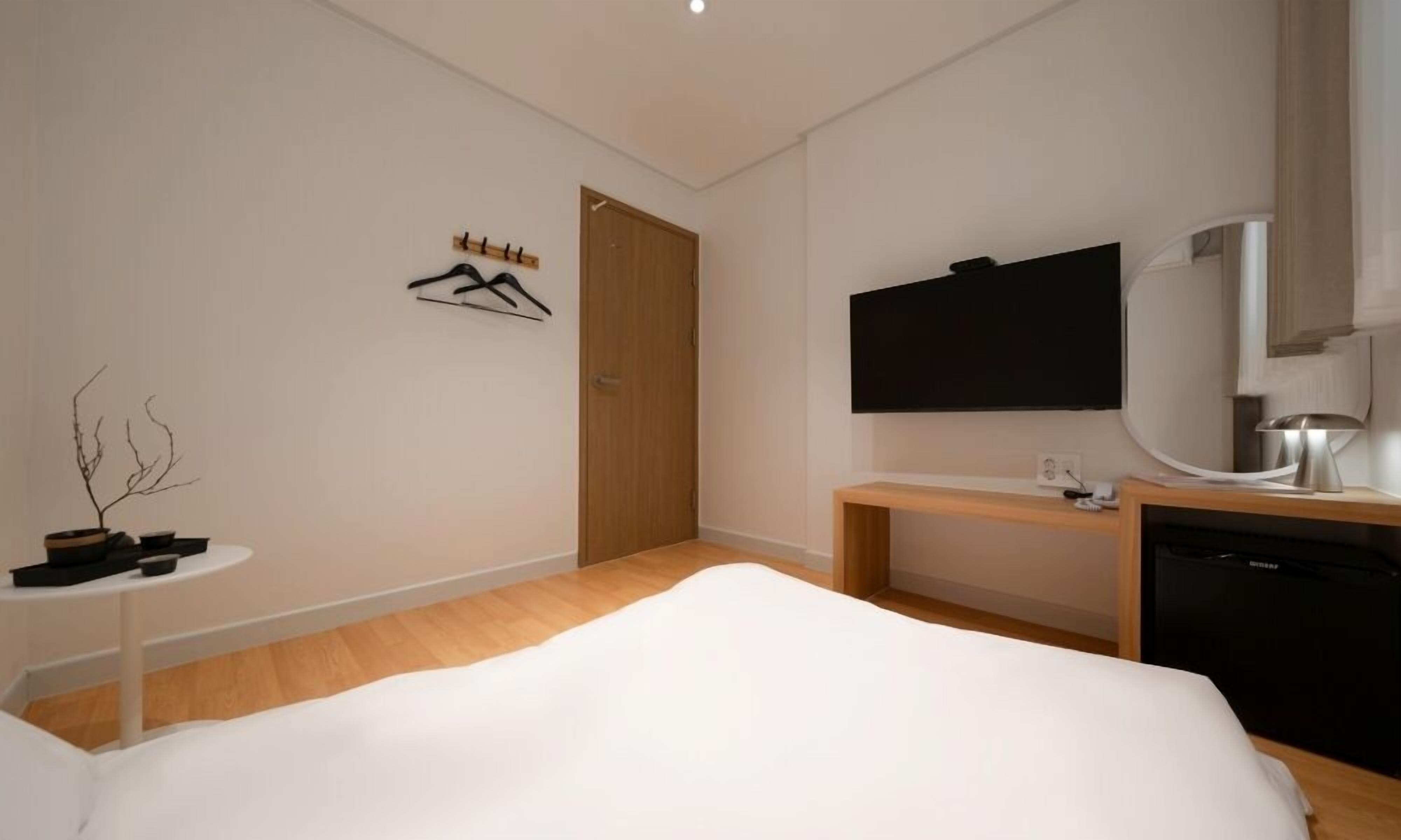Una habitación de hotel moderna con cama, televisor, escritorio con lámpara, una mesita con planta y una puerta de madera.