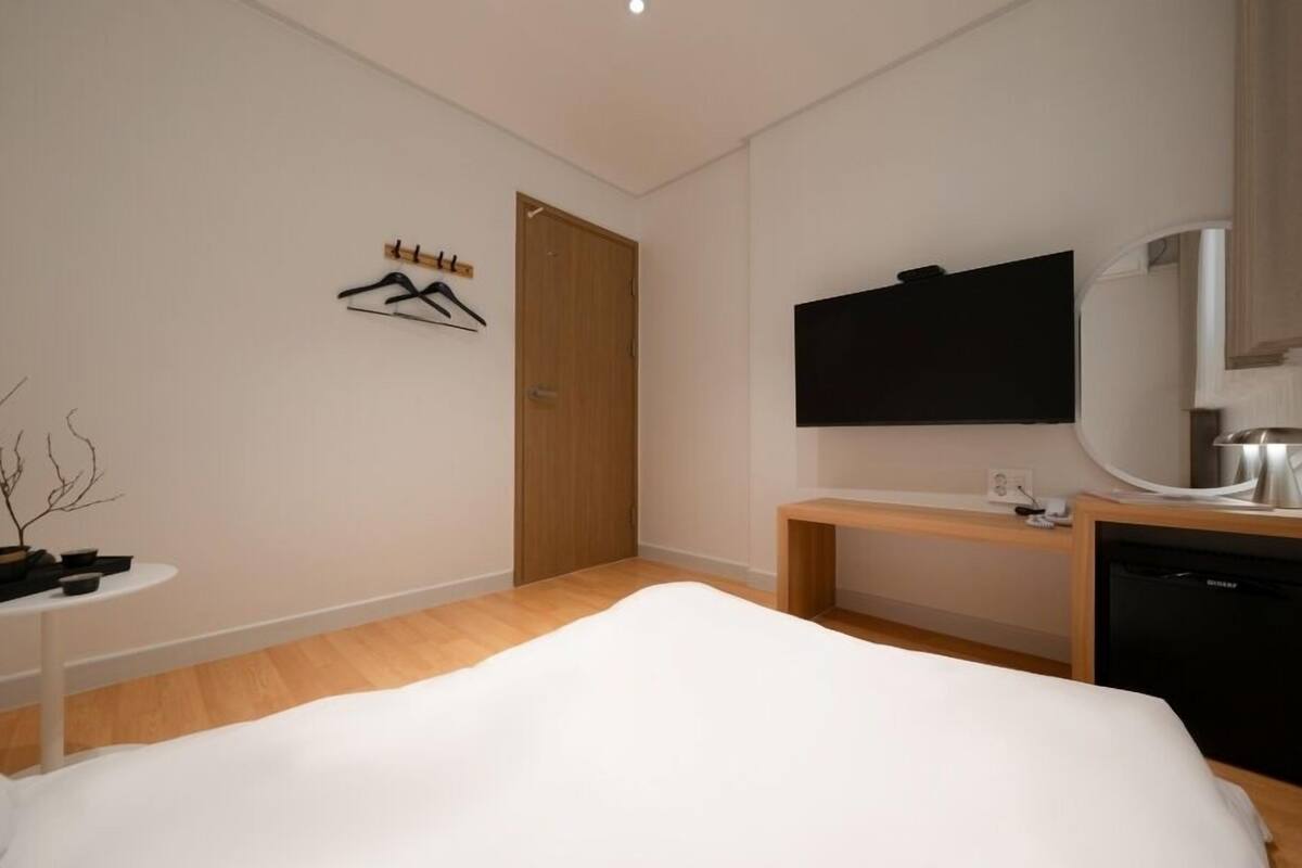 A modern hotel room with a bed, a TV, a desk with a lamp, a small table with a plant, and a wooden door.