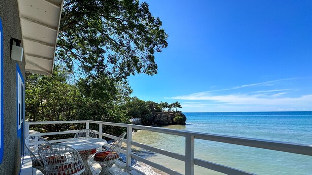 Eine Holzterrasse mit Sitzgelegenheiten im Freien, mit Blick auf das klare, blaue Meer und einen felsigen Landvorsprung mit Bäumen.